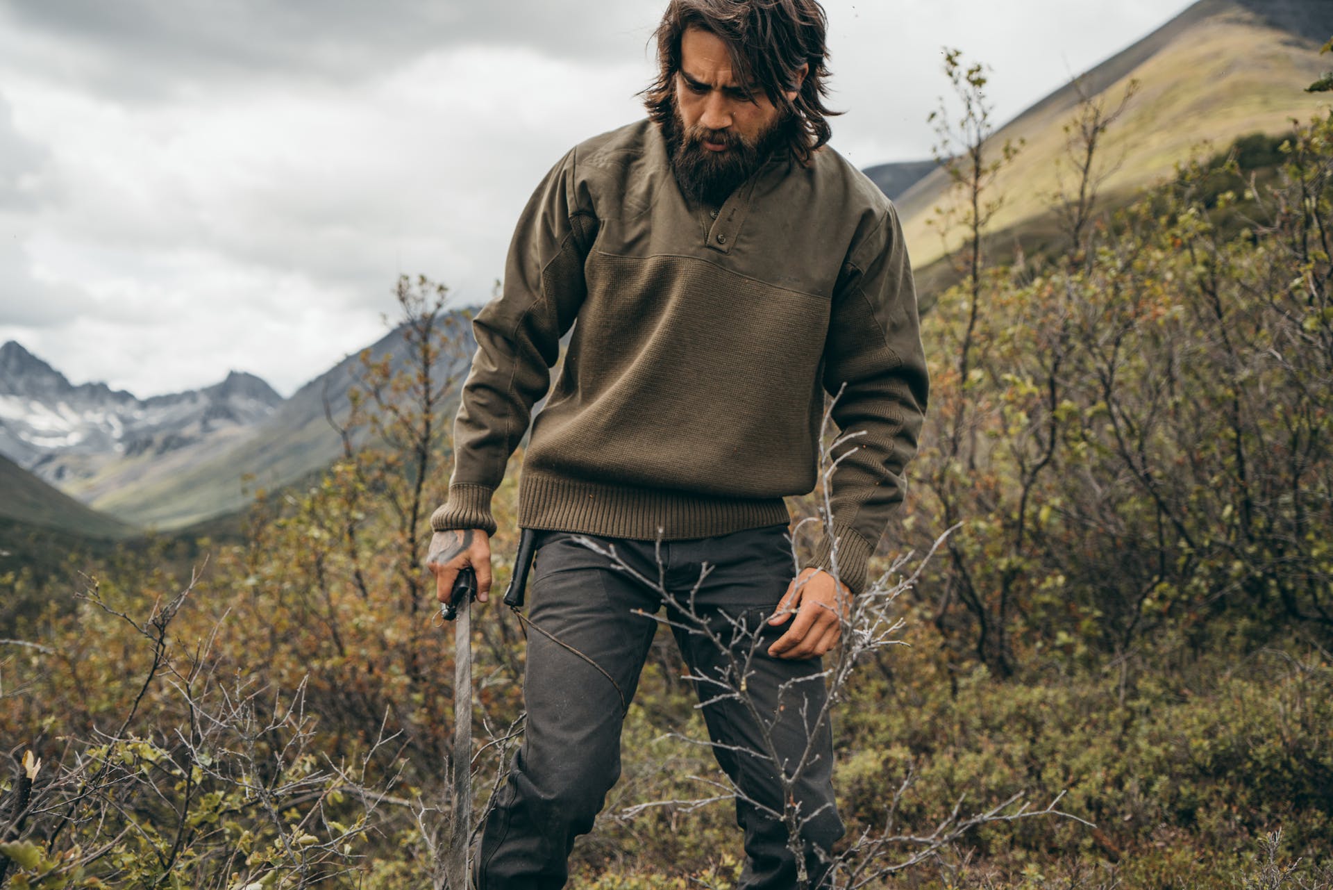 Man wearing Filson Henley Guide Sweater in peat green holding a machete and looking at shrub to cut