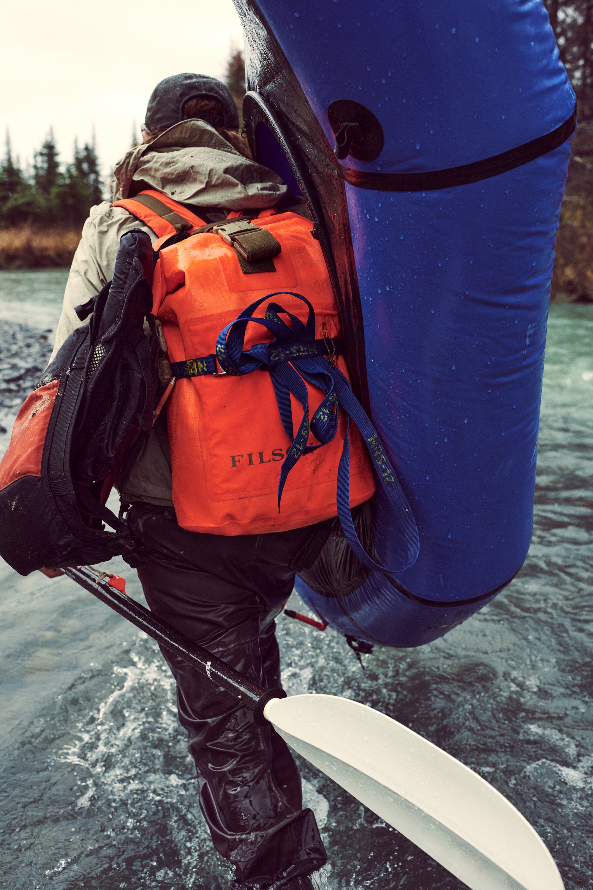 Man wearing Filson Dry Backpack in flame walking through water and holding rafting gear