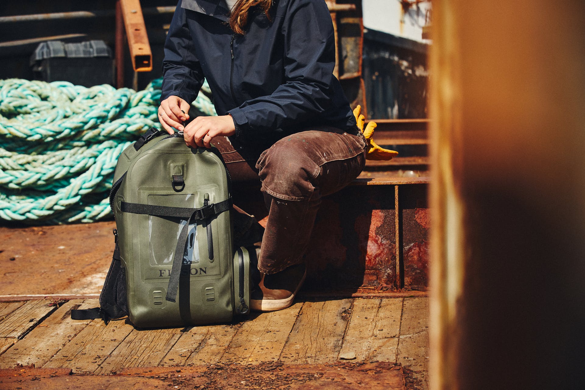 Woman sitting on a rusty old dock unzipping a Filson Backpack Dry Bag in green
