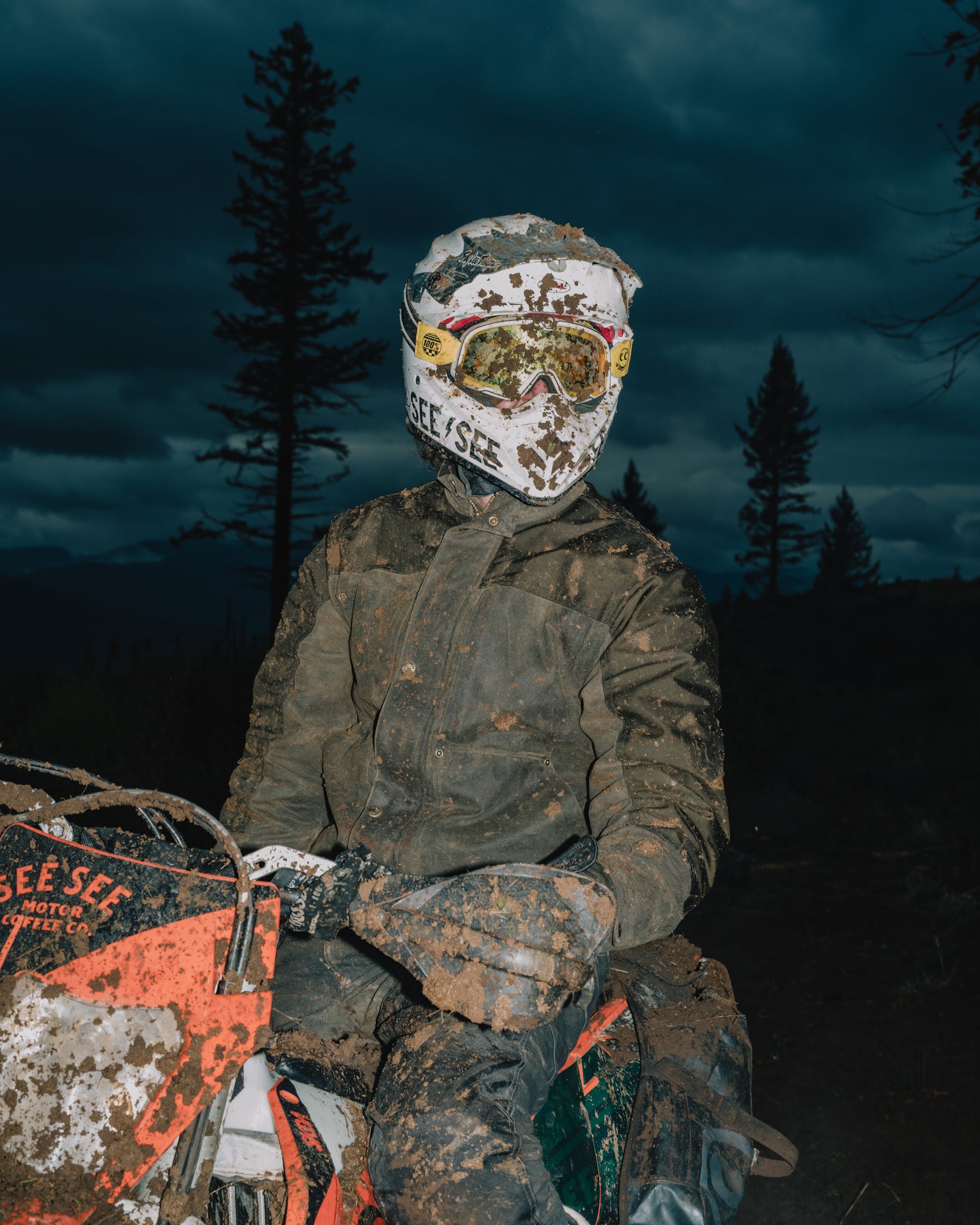 Man wearing Filson Alcan Canvas Cruiser in root covered in mud and sitting on motorcycle